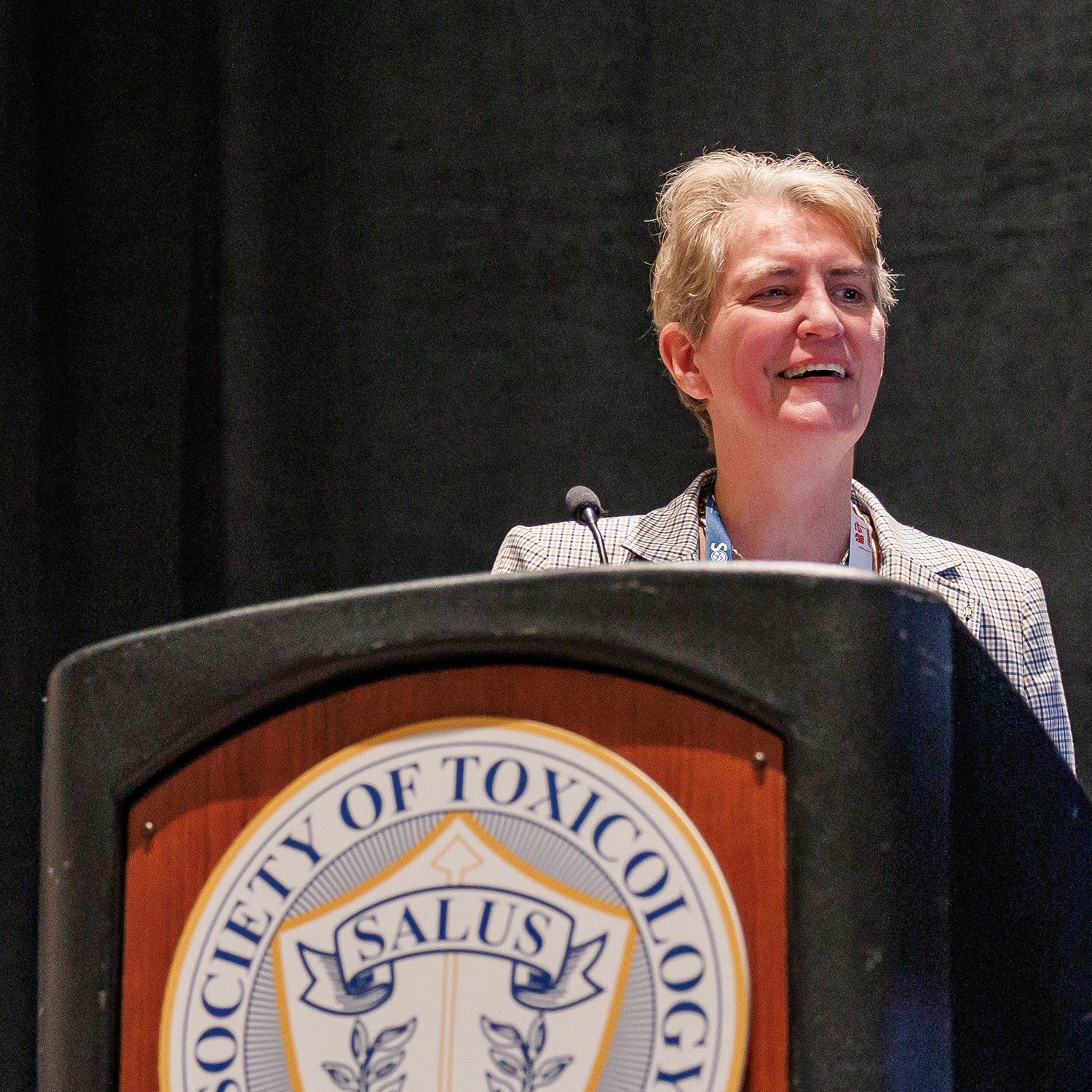 An individual is seated at a table. There is a microphone on a stand sitting on the table in front of them. They are speaking into the microphone while gesturing with their hands.
