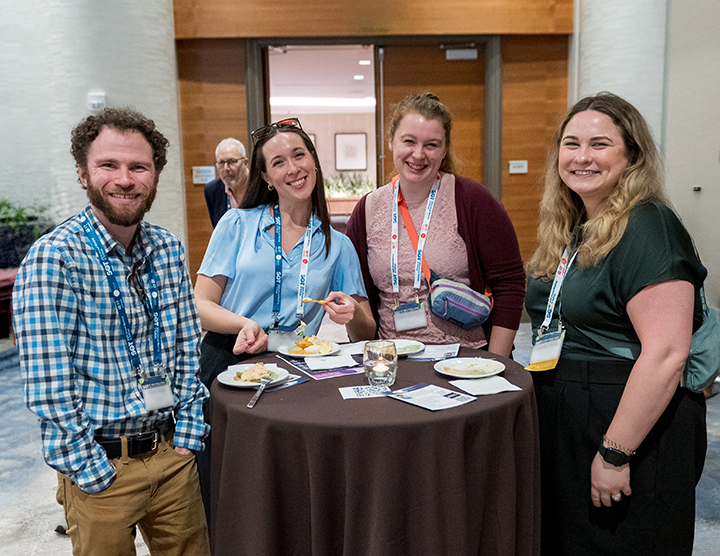 Four SOT Annual Meeting attendees smile together around a standing table. 