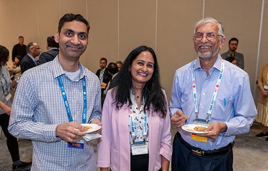 Three SOT Annual Meeting attendees smile for a photo, with other attendees networking in the background.